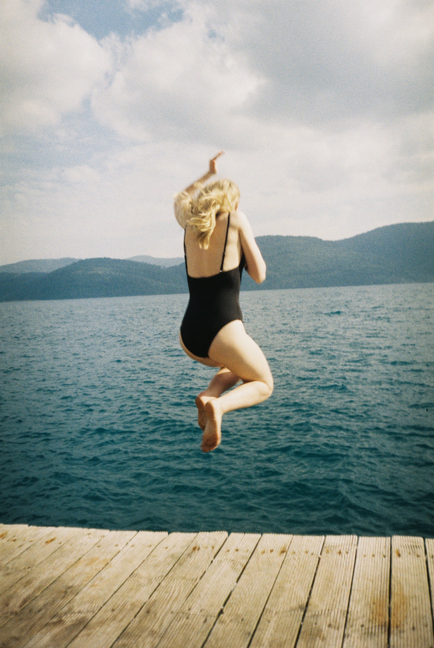 Person in a swimsuit jumping off a dock into a body of water with mountains in the background