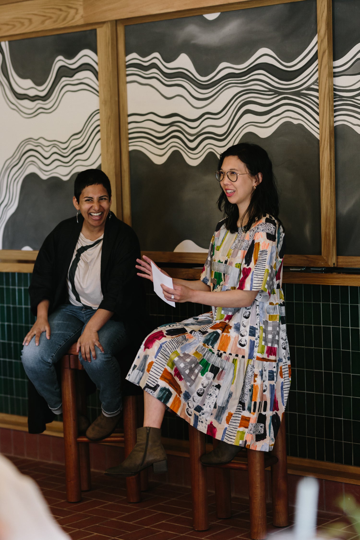 Two women sitting on stools in a room with decorative wall art.
